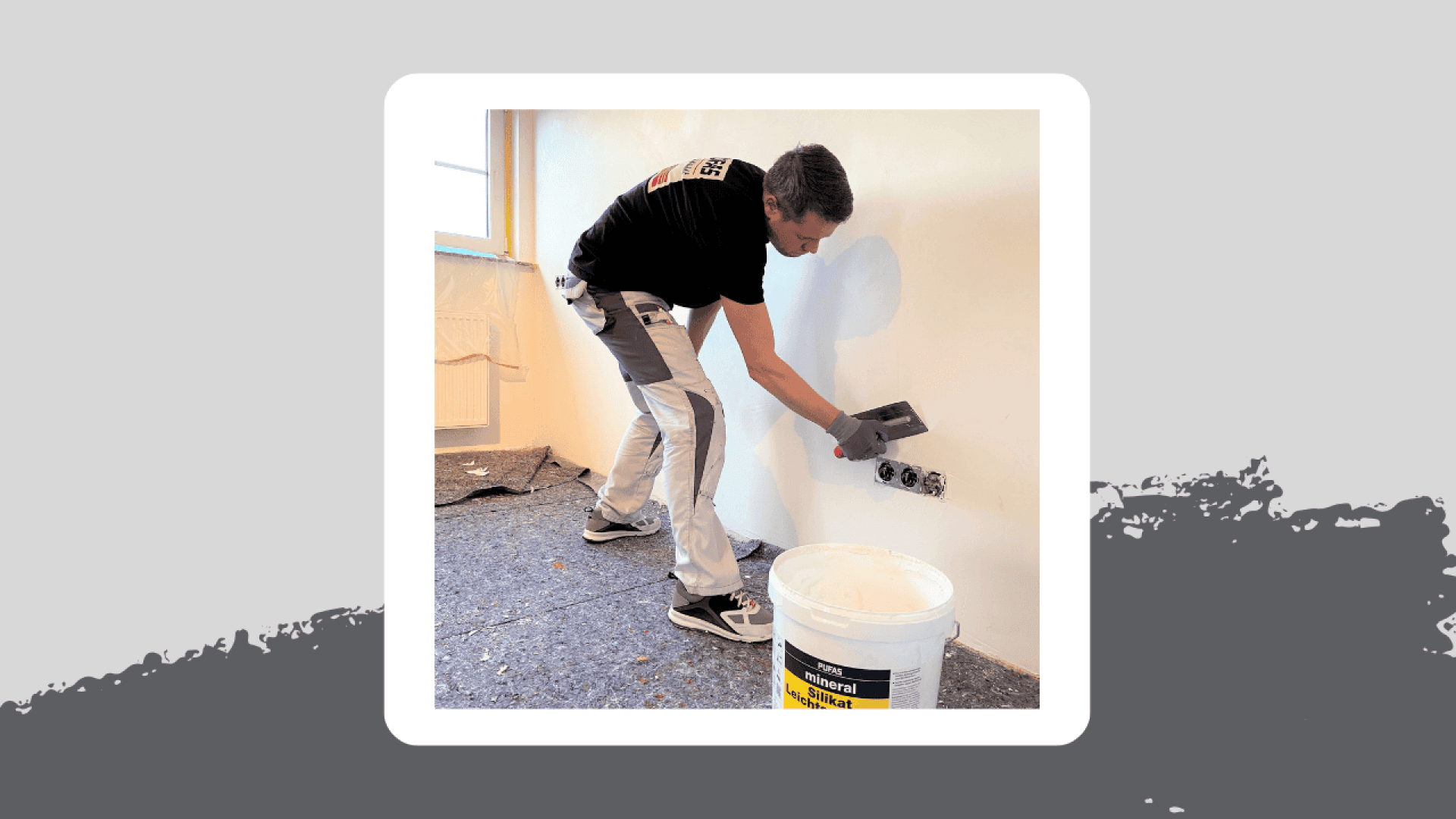 A person in work clothes uses a trowel to apply plaster to a white interior wall near electrical outlets. A white bucket of plaster sits on the floor nearby.