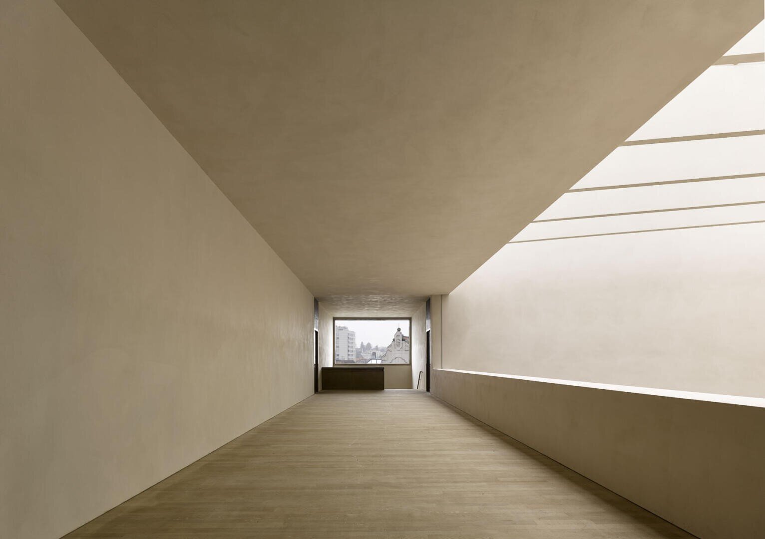 Ein minimalistischer, lichtdurchfluteter Flur mit glatten beigen Wänden, Holzboden, großem rechteckigem Oberlicht und einem entfernten Fenster mit Blick auf Dächer und eine Kirche im Hintergrund.