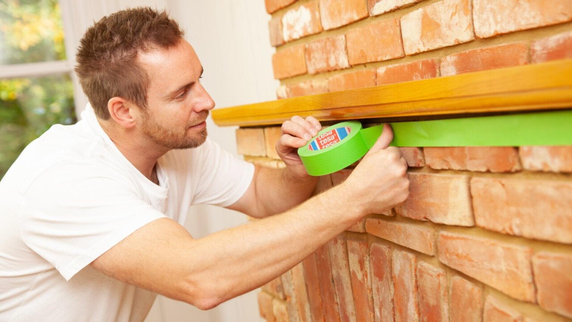 A man in a white shirt applies green painter’s tape to a brick wall beneath a wooden mantel, preparing the area for painting or another project.