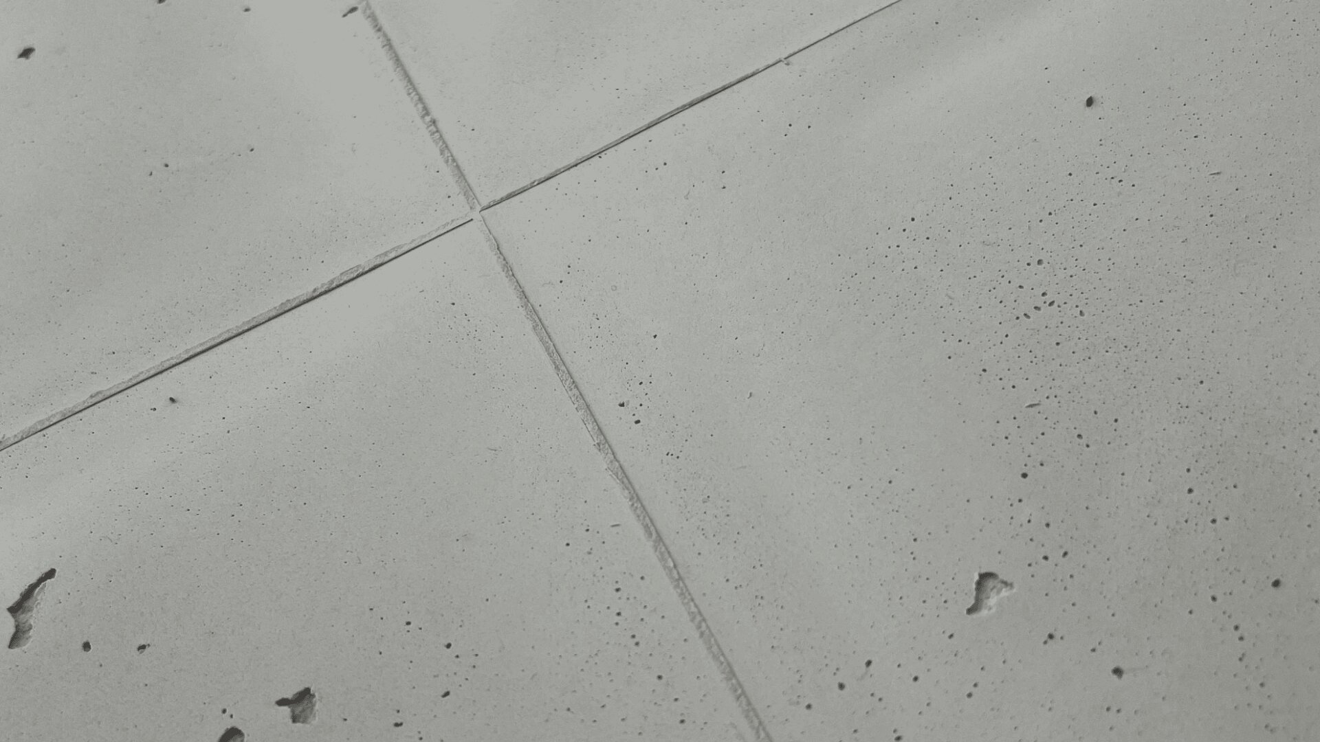 Close-up of a smooth concrete surface with intersecting lines forming a cross shape. The surface has small holes and imperfections, giving it a textured and industrial appearance.