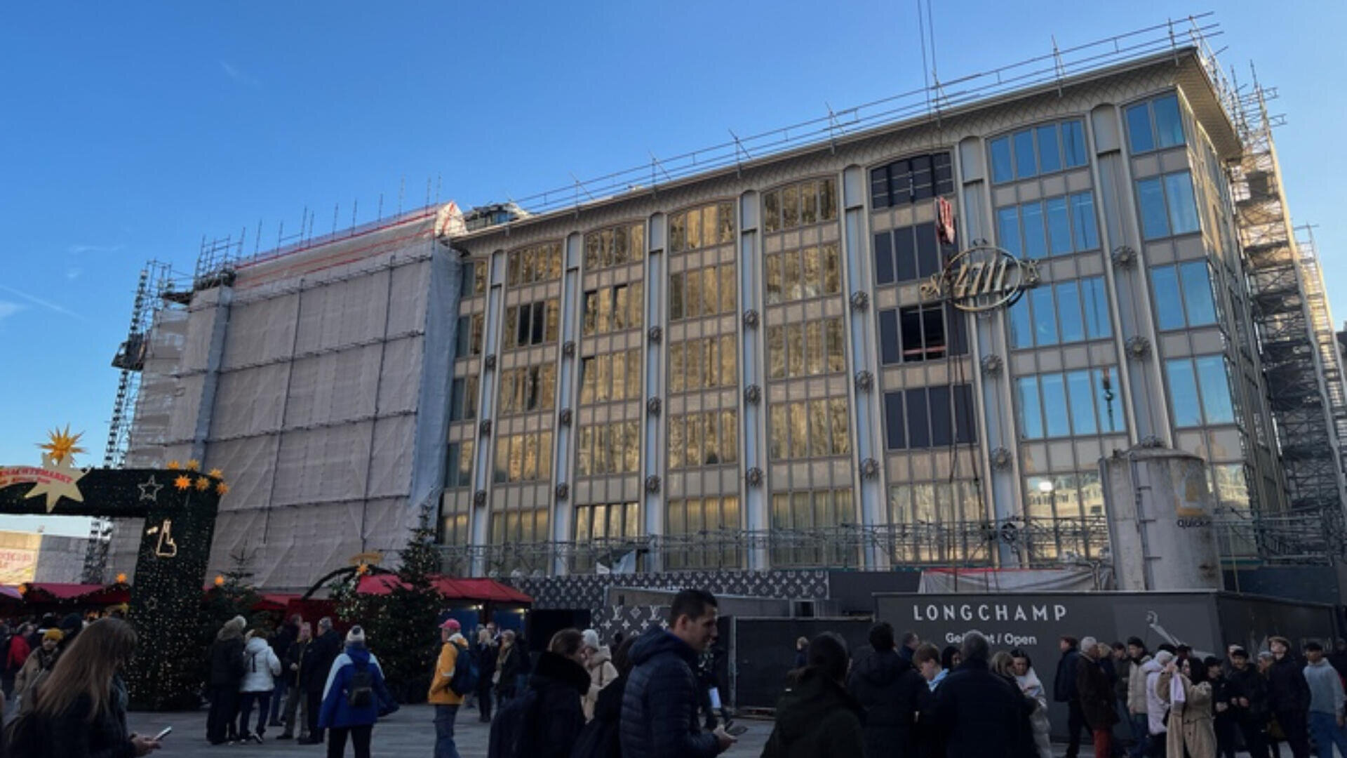 Ein großes Gebäude wird gerade renoviert. Gerüste und überdachte Bereiche reflektieren das Sonnenlicht. Menschen versammeln sich auf dem Platz davor. Marktstände, Weihnachtsbäume und festliche Dekorationen sind zu sehen. Der Himmel ist klar und blau.