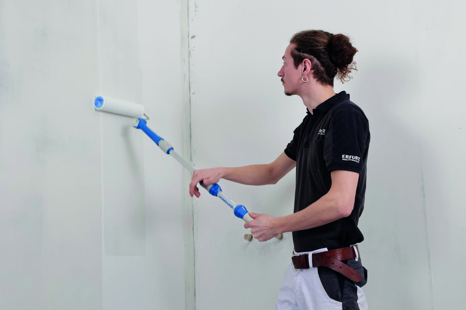Eine Person mit dunklem Hemd und heller Hose streicht mit einer Farbrolle an einer Verlängerungsstange eine weiße Wand.