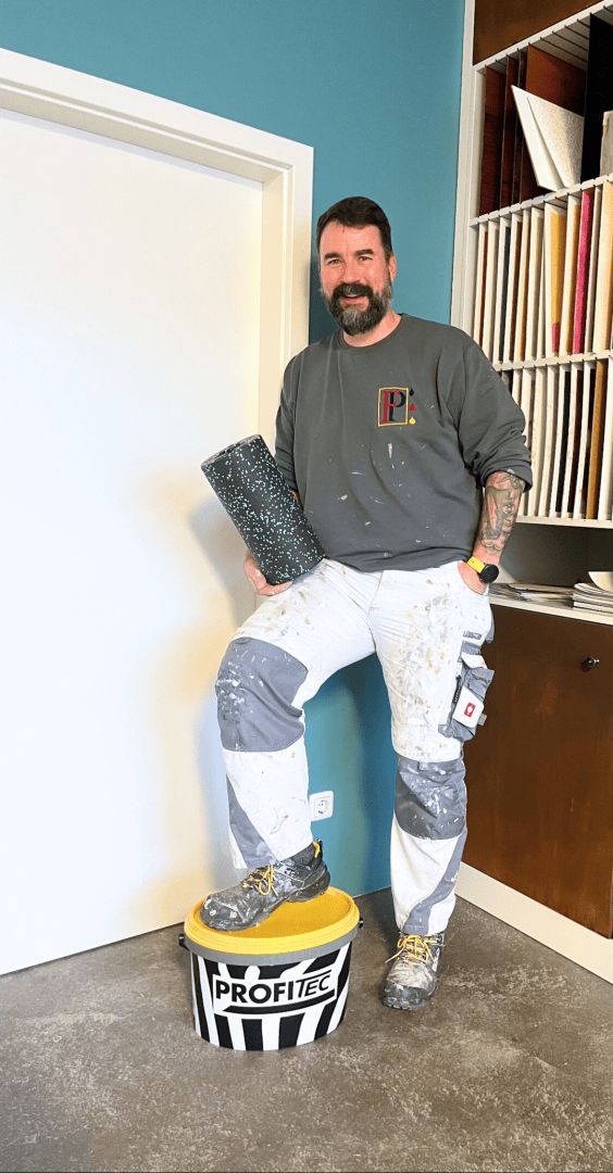 Ein bärtiger Mann in Arbeitskleidung steht lächelnd im Innenbereich, einen Fuß auf einem PROFITEC-Eimer und hält eine Rolle Bodenbelag oder Material in der Hand. Er steht vor einer weißen Tür und einer blauen Wand.