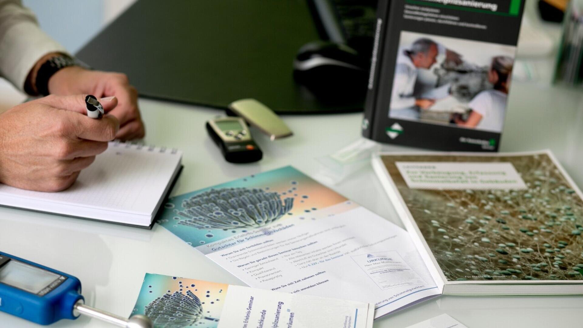 A person writes in a notebook on a desk with brochures, documents, a book, a car key, and a device. The materials appear to relate to science or medicine, featuring images of cells and scientific illustrations.