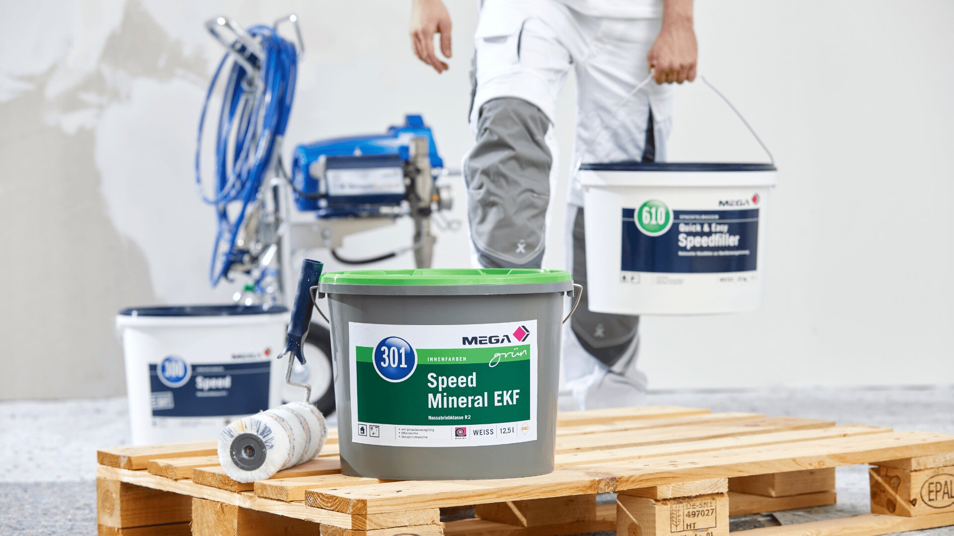 Eine Person in weißer Arbeitskleidung und grauen Knieschonern steht auf einer Holzpalette und hält einen Eimer mit Wandspachtel. Im Hintergrund stehen Eimer mit Wandspachtel und ein Farbspritzgerät.