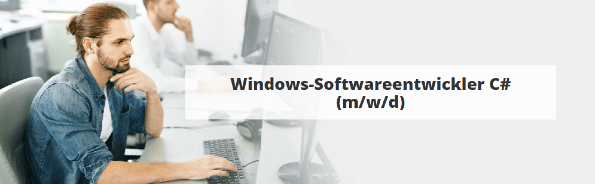 Ein Mann mit Bart und zurückgebundener Frisur arbeitet an einem Computer in einem Büro. Der Text auf dem Bild lautet: Windows-Softwareentwickler C# (m/w/d).