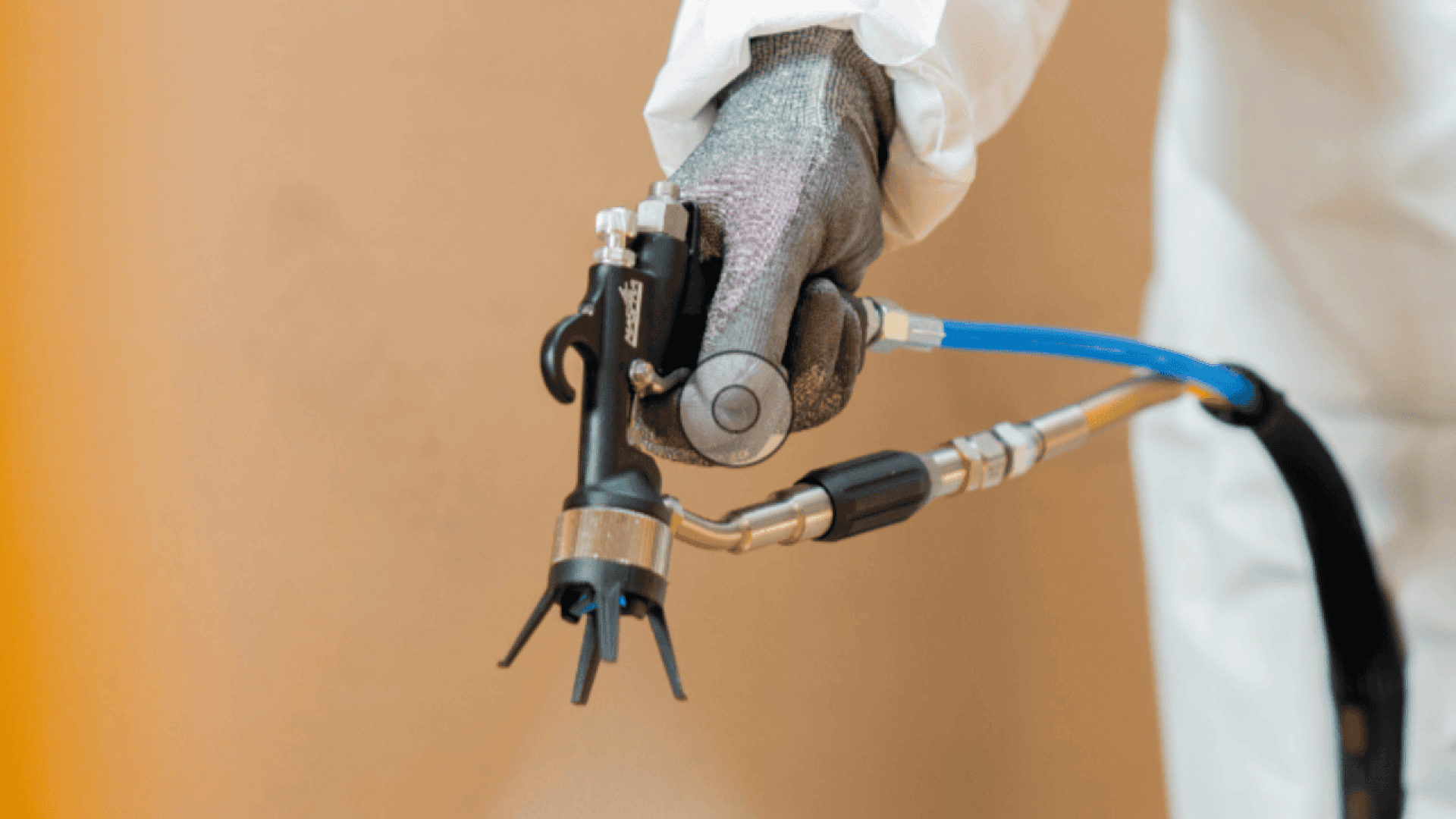 A gloved hand holds a pesticide sprayer with blue and black hoses, wearing protective white clothing, against a plain beige background.