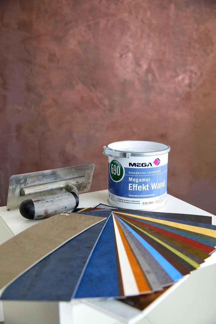 Eine Farbdose mit der Aufschrift „MEGA Megamur Effekt Wand“, eine Kelle mit Rolle und eine aufgefächerte Auswahl an Farbmustern werden auf einer weißen Fläche vor einer strukturierten, braun gestrichenen Wand präsentiert.