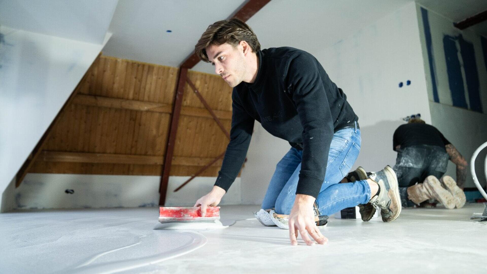 A man in a black sweater and jeans kneels on the floor, smoothing a white surface with a red tool. Another person works in the background. The room has slanted wooden beams and unfinished walls.
