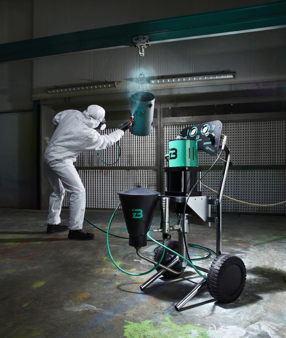 Eine Person in Schutzkleidung lackiert ein von der Decke hängendes Metallobjekt. Im Vordergrund ist auf dem Werkstattboden eine industrielle Farbspritzmaschine mit Schläuchen und Rädern zu sehen.