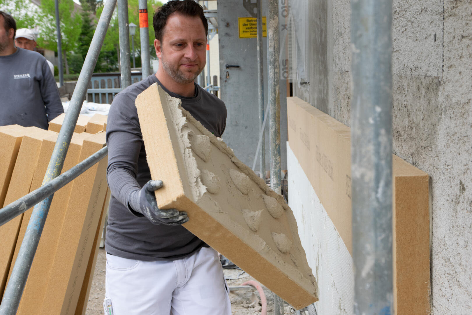 Ein Bauarbeiter hält eine Dämmplatte mit Klebstoff in der Hand und bereitet sie vor, um sie auf einer Baustelle an einer Außenwand zu befestigen. Im Hintergrund sind ein weiterer Arbeiter und weitere Dämmplatten zu sehen.