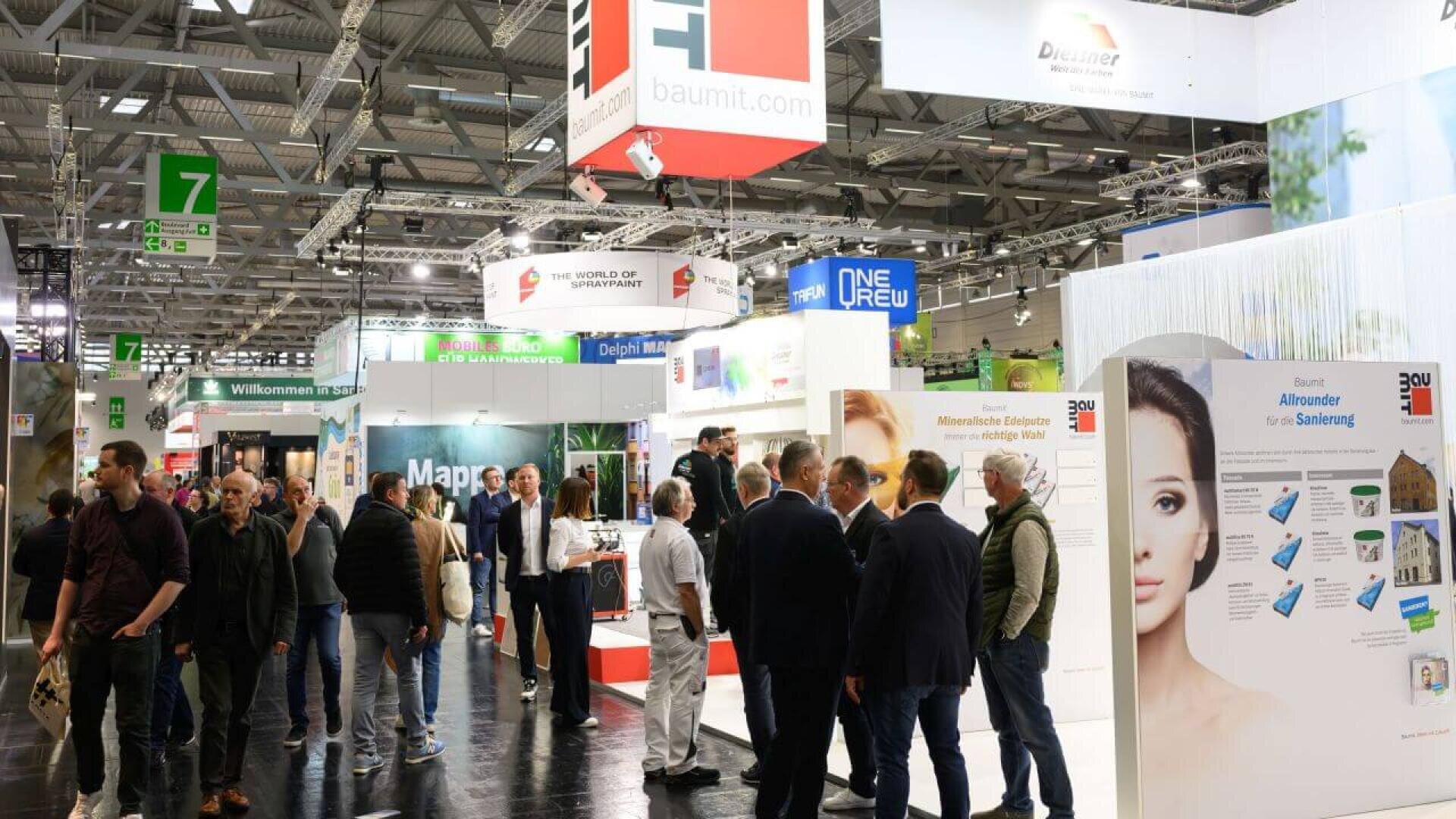 Eine geschäftige Messe mit vielen Menschen, die zwischen den verschiedenen farbenfrohen Ständen und Ausstellungen, darunter auch prominente Schilder von Baumit und anderen Unternehmen, umhergehen und sich unterhalten. Die Umgebung ist mit Deckenleuchten und Bannern gut beleuchtet.