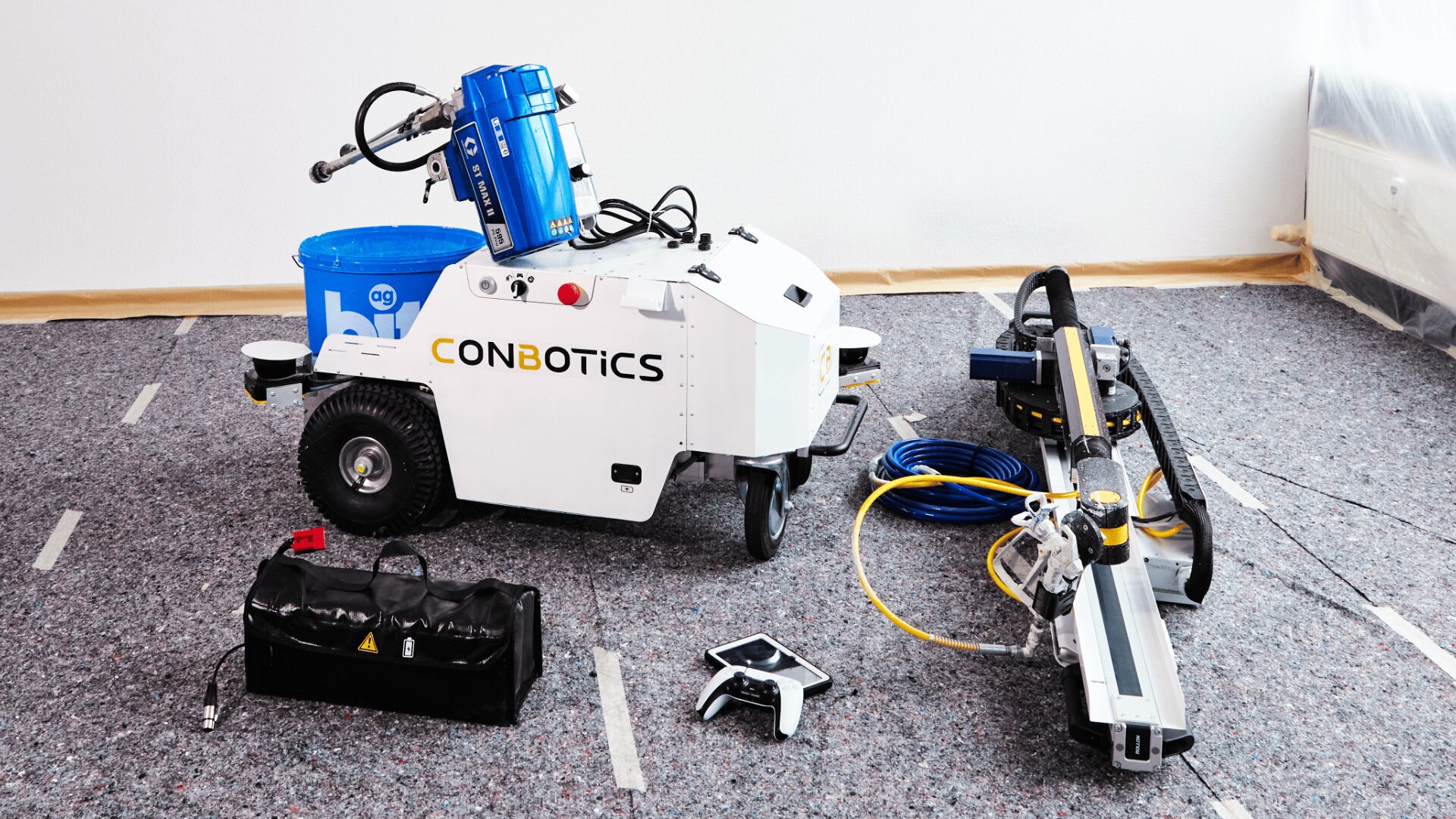 A white robot labeled CONBOTICS is on a carpeted floor, surrounded by a blue bucket, a tool bag, a control device, electrical cables, and a metal machine attachment in a bright indoor setting.