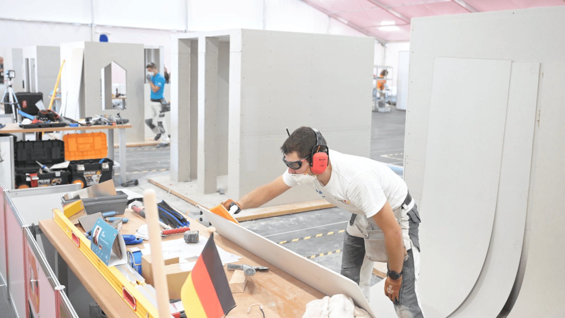 Eine Person in Schutzkleidung arbeitet in einem Gebäude an einer Baustelle und hantiert mit einer großen Gipskartonplatte. Werkzeuge und eine deutsche Flagge liegen auf einem Tisch daneben. Im Hintergrund sind unfertige Wände zu sehen.