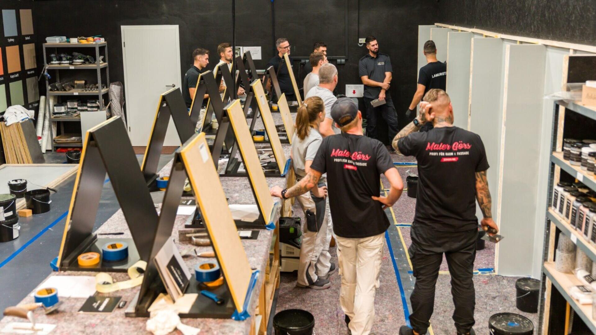A group of people in casual workwear gather in a workshop, surrounded by painting stations and supplies, listening to an instructor near partially painted boards and shelves of materials.