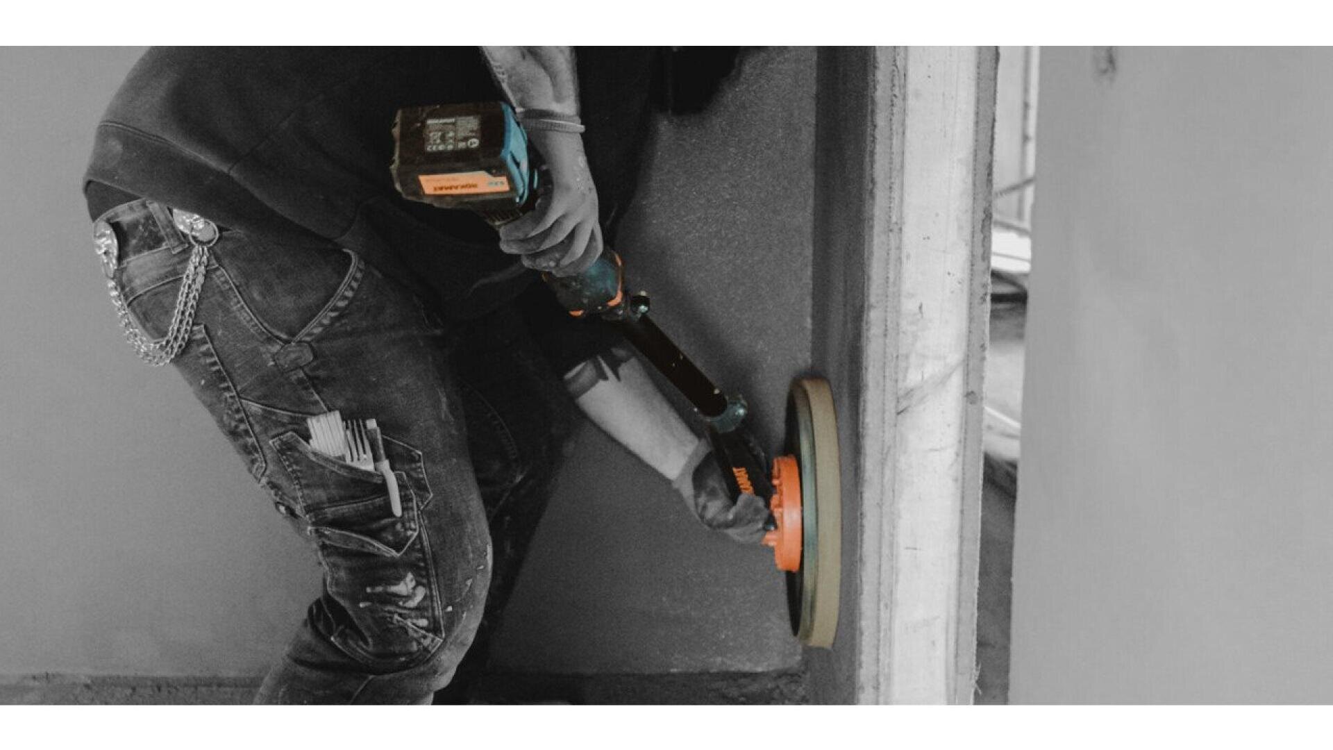A person using a power tool to sand or polish a wall. The person is wearing jeans with tools in the pocket and a chain hanging from the belt loop. The image is mostly black and white, with the tool highlighted in color.