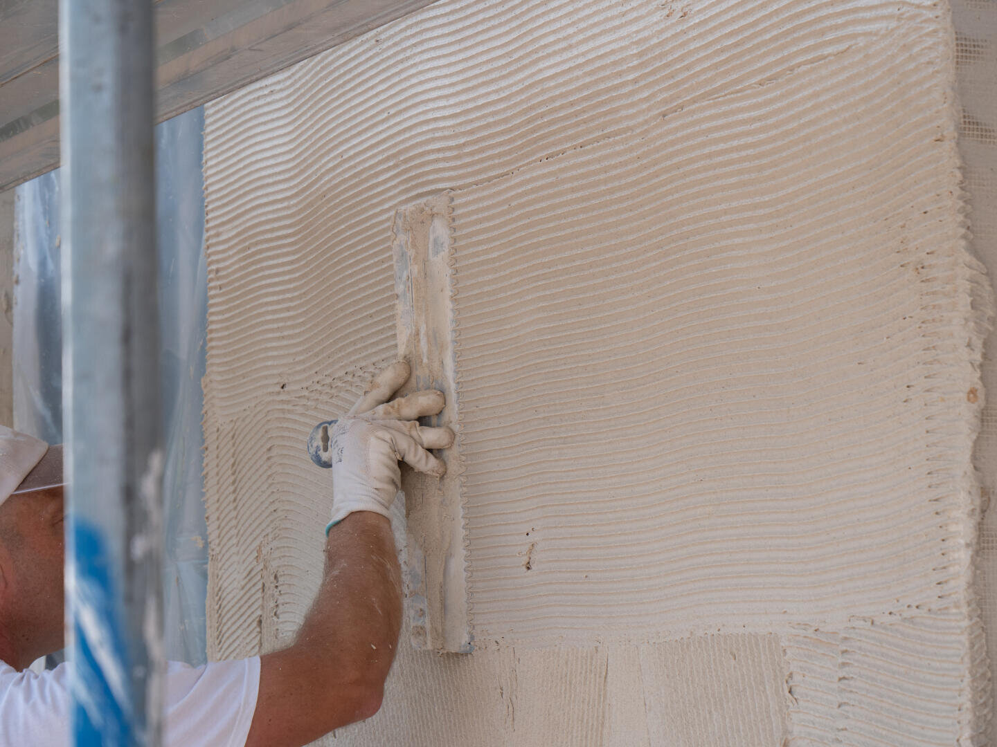 Eine Person mit einem weißen Handschuh glättet mit einer Kelle den Putz einer Wand und sorgt so bei Bau- oder Renovierungsarbeiten für eine gleichmäßig strukturierte Oberfläche.