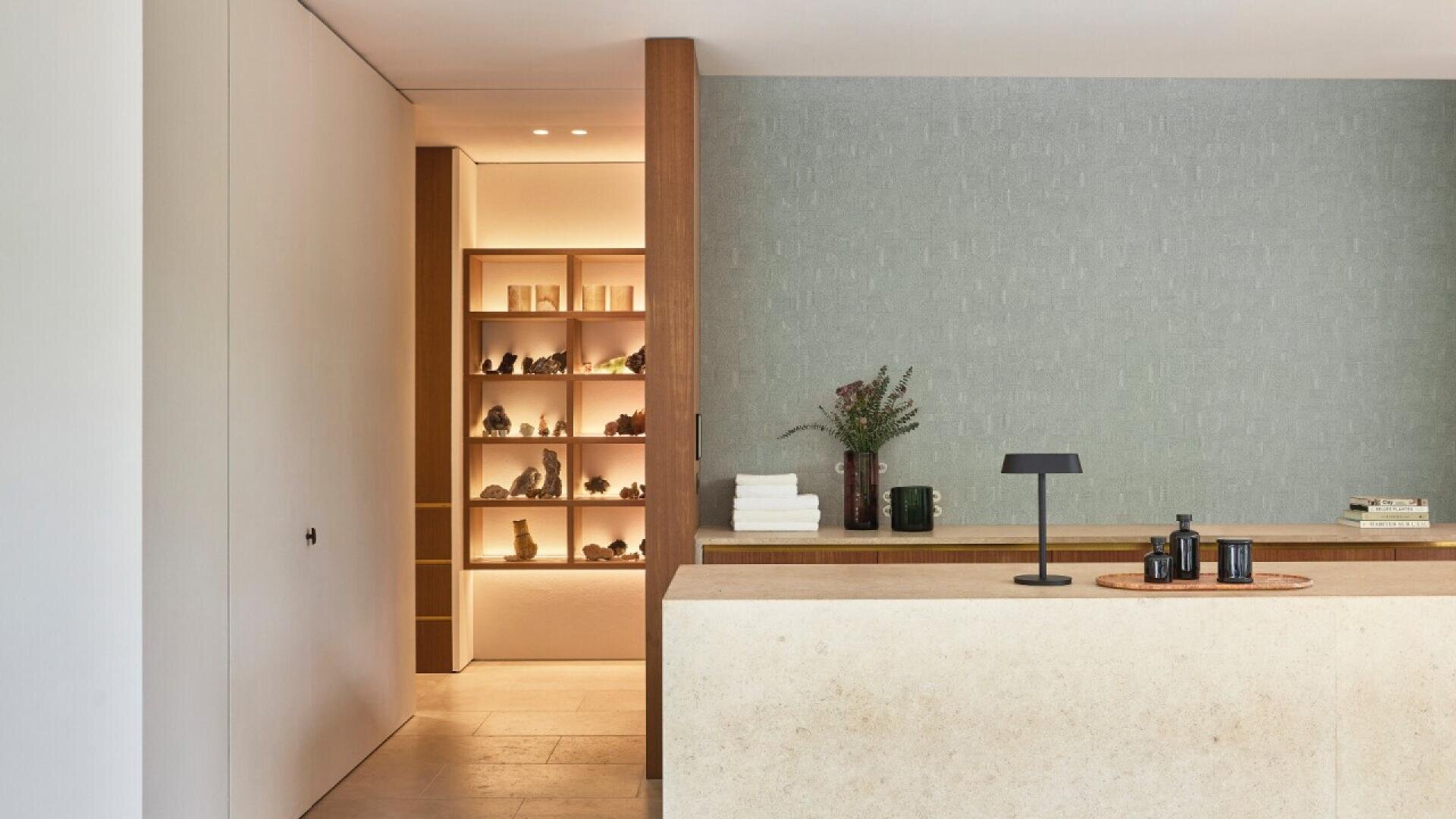 Minimalist reception area with a stone desk, a small black lamp, stacked towels, flowers in a vase, and decorative items on a wooden tray. In the background, a softly lit shelf displays various objects.