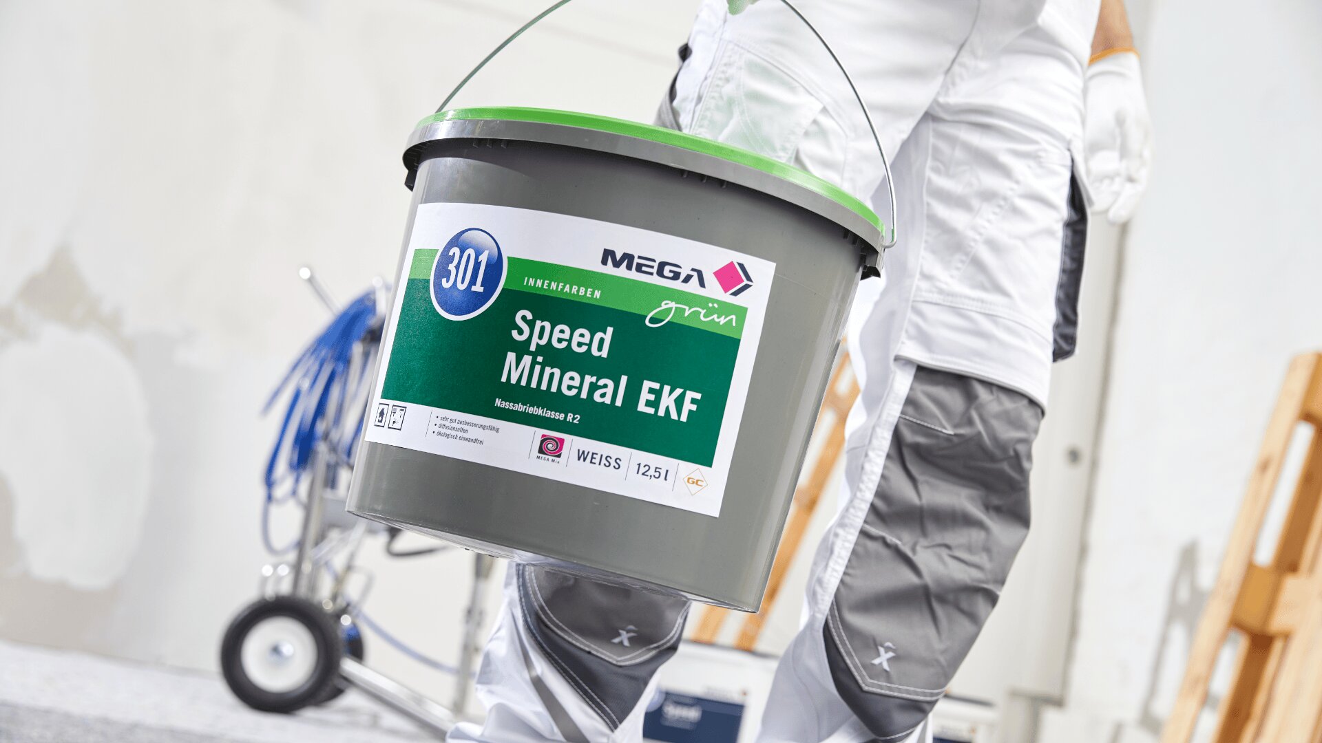 Eine Person in weißer Arbeitskleidung und grauen Knieschützern hält einen grünen Eimer mit der Aufschrift „MEGA Speed Mineral EKF“ in einer Innenumgebung mit Malutensilien und einer Leiter im Hintergrund.