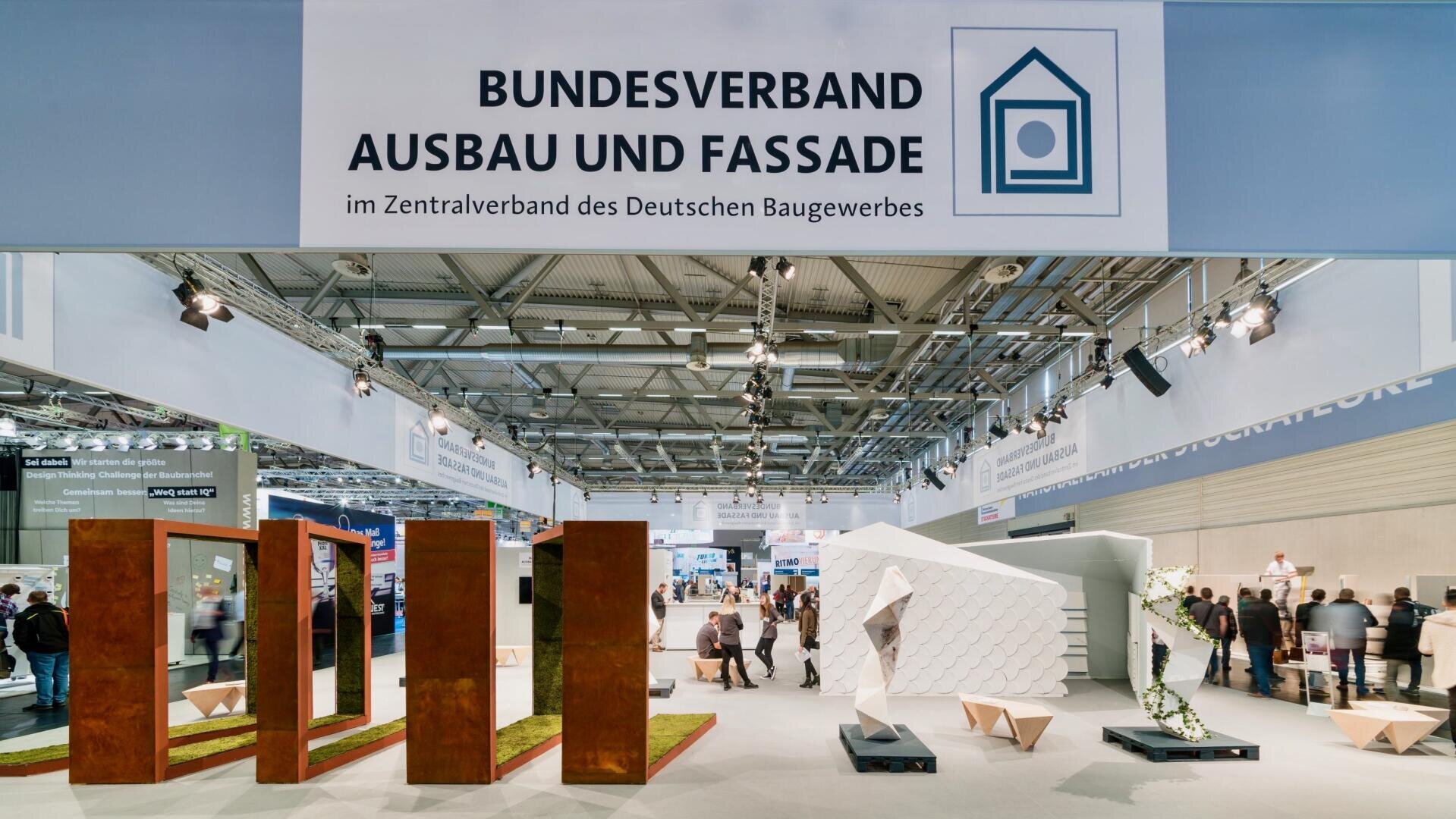 Messestand des Bundesverbandes Ausbau und Fassade mit modernen Baudisplays, Spiegeln und weißen geometrischen Strukturen; Menschen laufen in einer großen, hellen Messehalle umher.