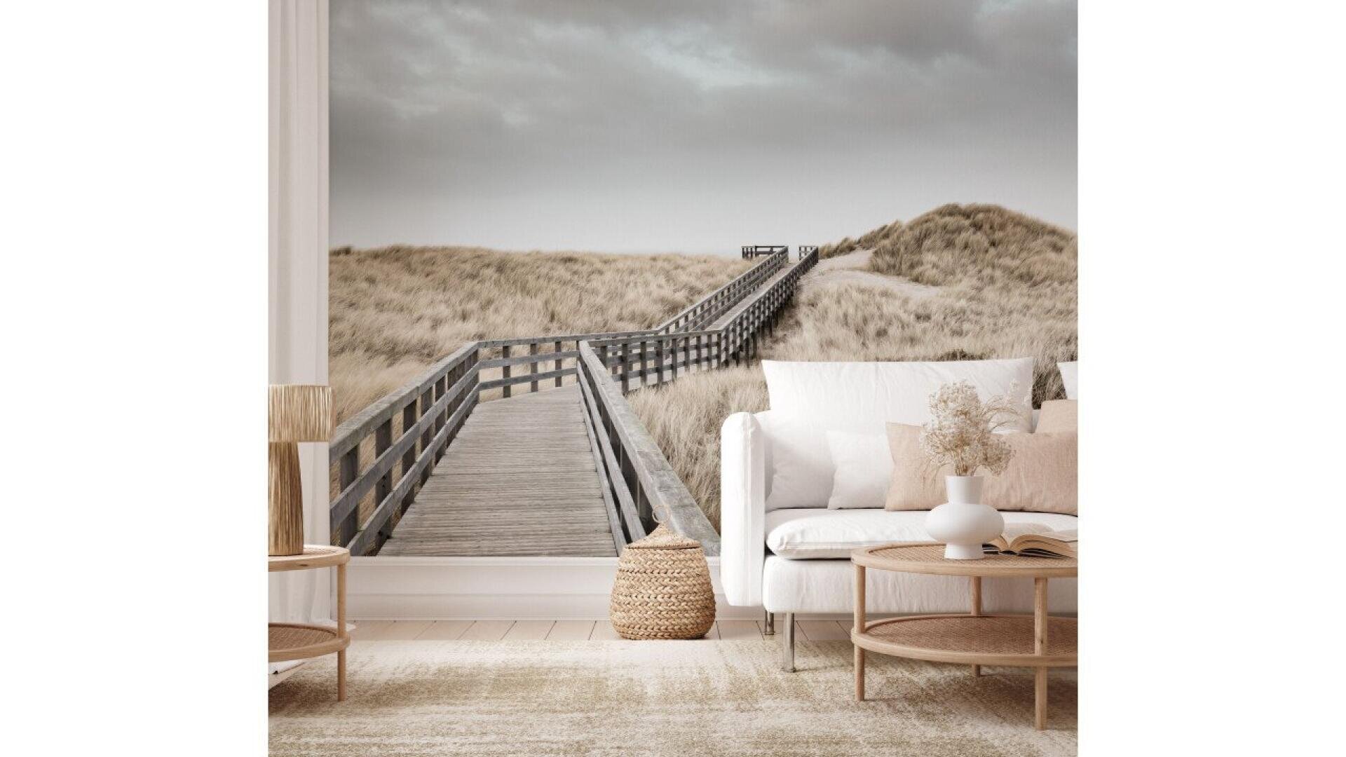 A cozy living room with neutral decor and light furniture, featuring a large mural of a wooden boardwalk leading through sand dunes under a cloudy sky as the main wall backdrop.