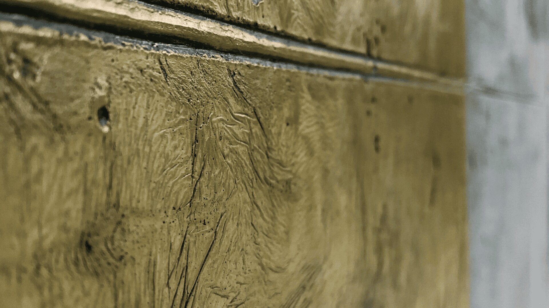 Close-up view of a textured, gold-colored wall with visible brush strokes and grooves, divided by a horizontal groove, and a blurred gray section on the right side.