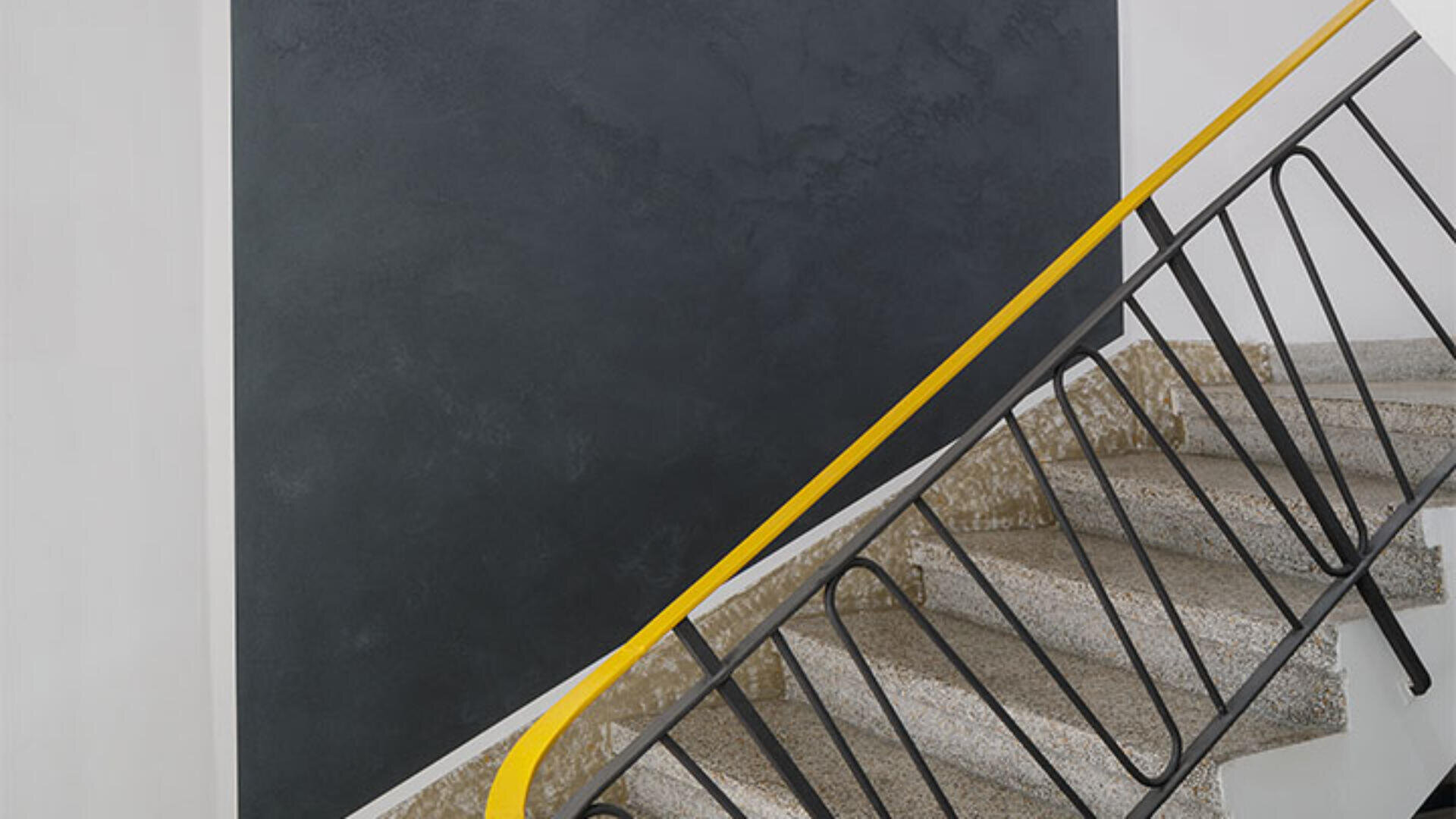 Eine Treppe mit Terrazzostufen und einem schwarzen Metallgeländer mit leuchtend gelbem Handlauf vor einer schlichten schwarzen Wand.