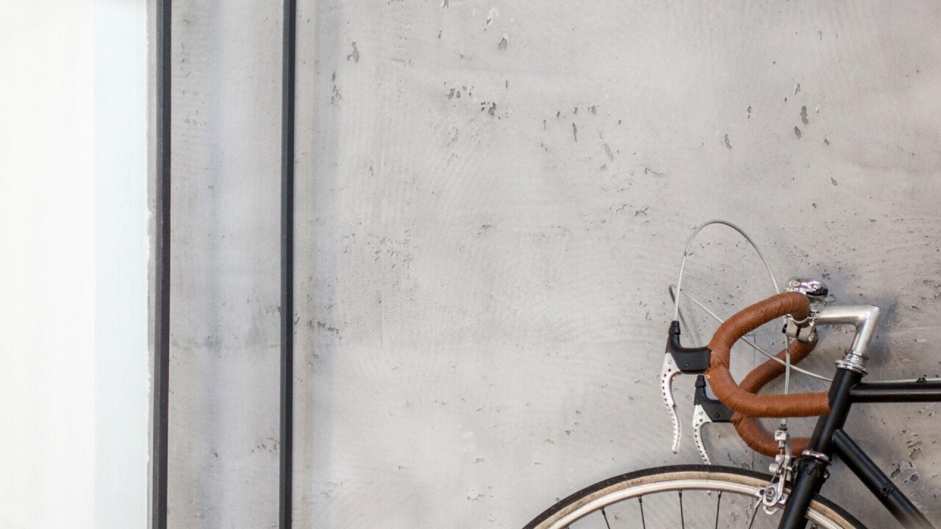 A close-up of a bicycle’s handlebars and front wheel, with brown handlebar tape, leaning against a light gray, textured concrete wall.