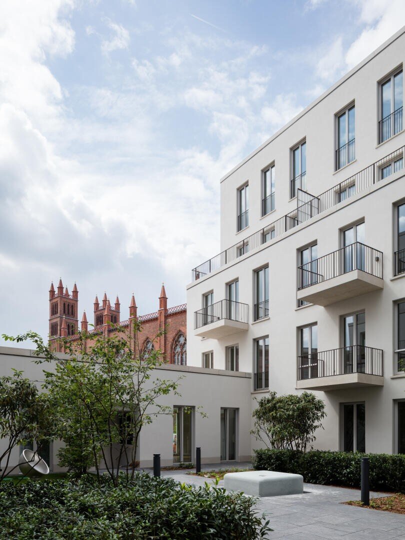 A modern white apartment building with balconies stands beside a courtyard with plants, set against a backdrop of a historic red brick church with tall spires under a partly cloudy sky.