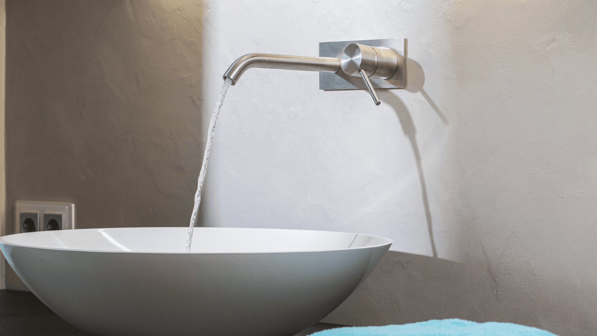 Ein modernes Badezimmerwaschbecken mit rundem, weißem Becken und einem eleganten, an der Wand montierten Edelstahlhahn, aus dem Wasser fließt. Im Hintergrund ist eine glatte, helle Wand mit einer sichtbaren Steckdose zu sehen.