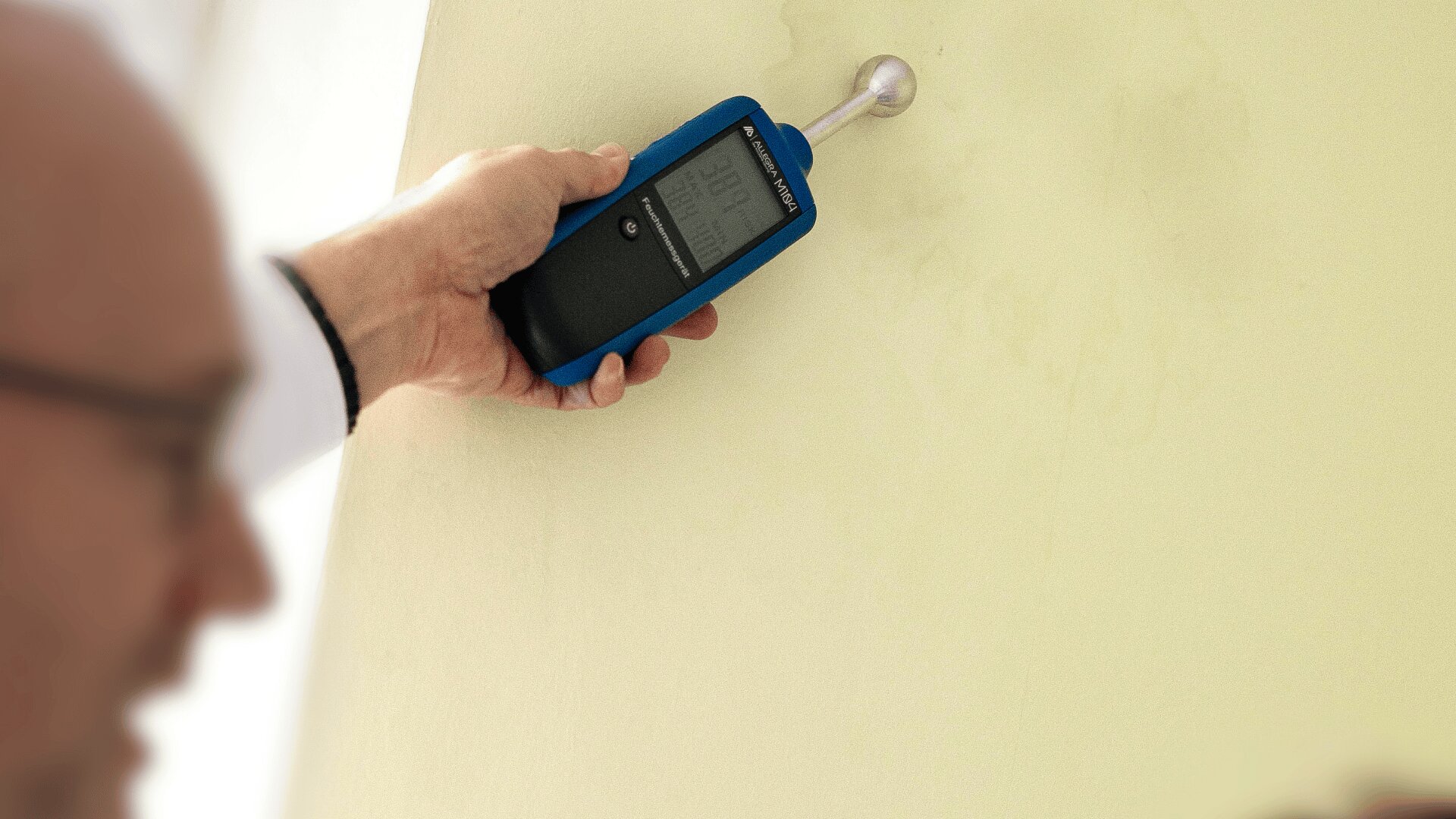 Eine Person hält ein digitales Feuchtigkeitsmessgerät an eine helle Wand, um den Feuchtigkeitsgehalt zu messen. Das Display des Geräts zeigt numerische Werte an. Das Gesicht der Person ist im Vordergrund unscharf dargestellt.