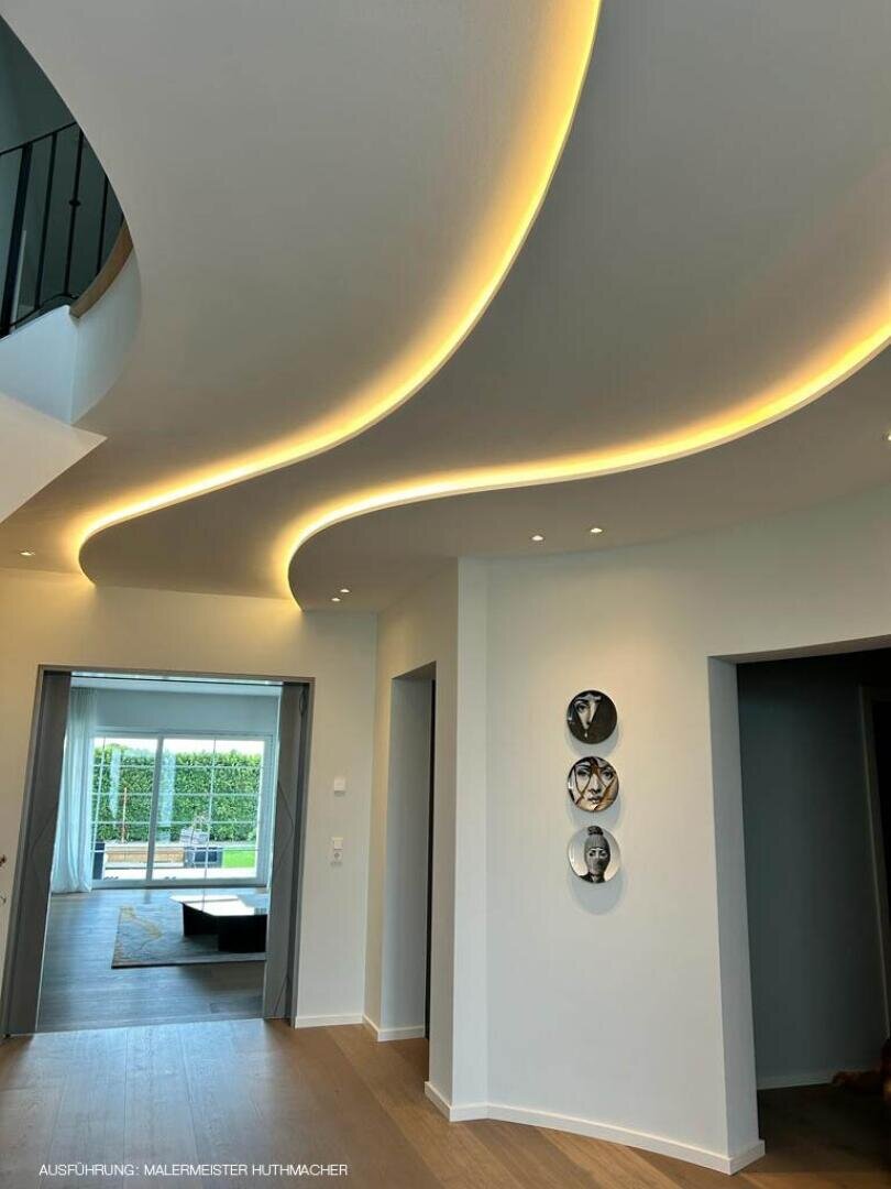 Ein moderner Flur mit gewellter, abgehängter Decke und warmer, indirekter LED-Beleuchtung. Drei Schwarz-Weiß-Porträts hängen an einer weißen Wand, und große Glastüren öffnen sich zu einer Terrasse und einer grünen Außenanlage.