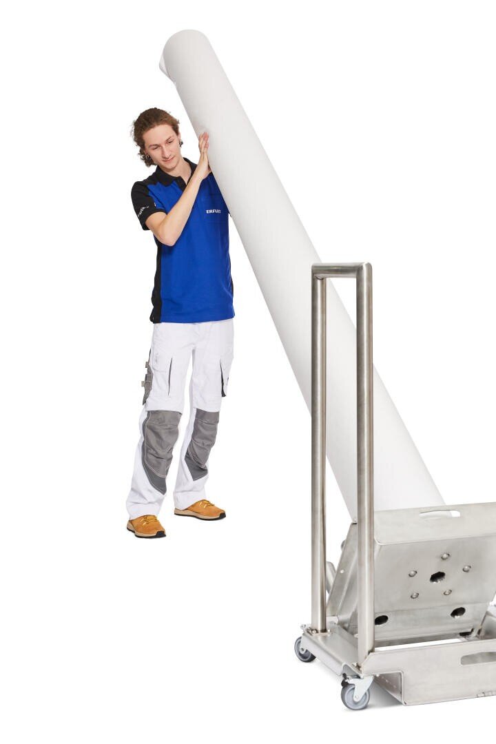Eine Person in einem blau-schwarzen Hemd und Arbeitshosen steht vor einem weißen Hintergrund neben einer großen, aufrecht stehenden Rolle weißen Materials auf einem Metallwagen mit Rädern.