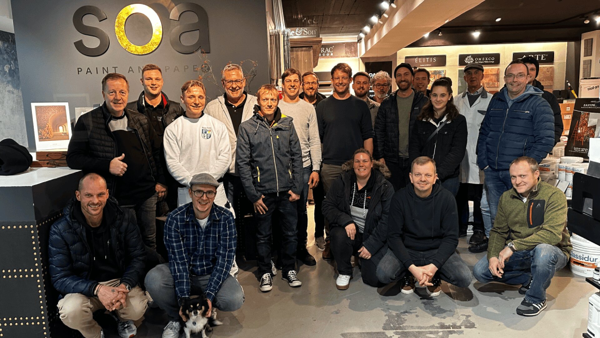 A group of around 20 people, mostly men, pose and smile for a photo inside a modern showroom with “soa paint and paper” on the wall behind them. Some people are standing, while others are kneeling at the front.