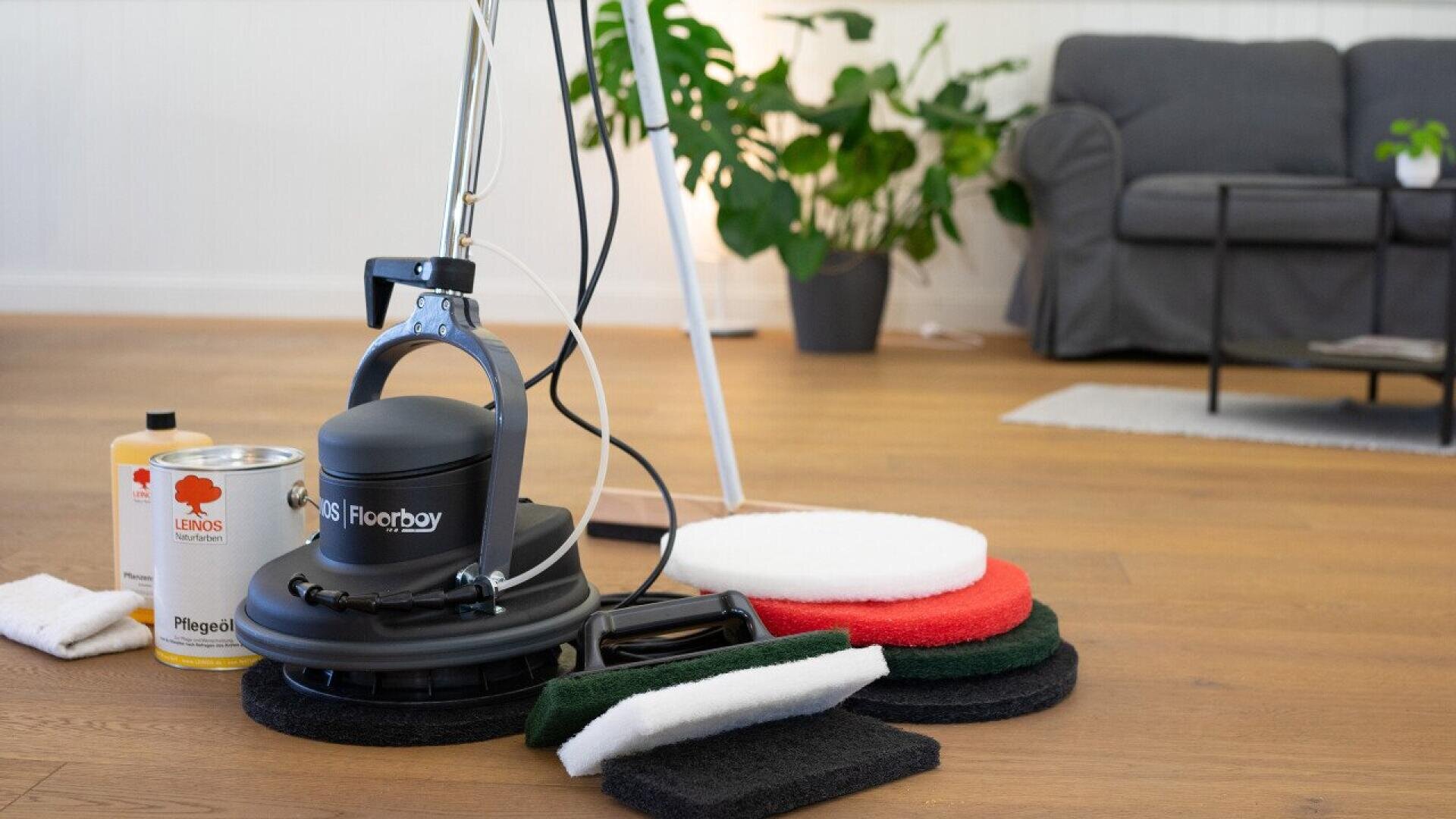 A floor cleaning machine with multiple colored pads and a bottle of cleaning solution sits on a wooden floor in a bright living room with plants and a gray couch in the background.