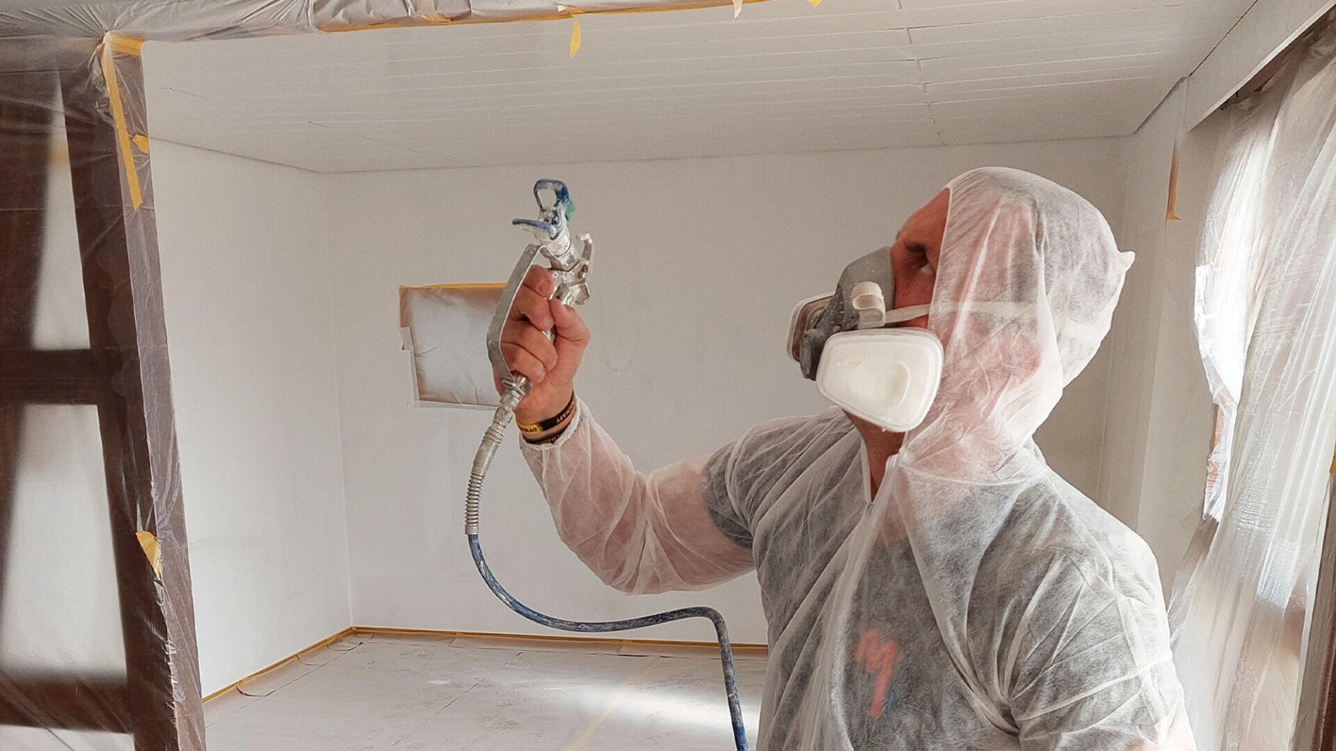Eine Person in Schutzanzug und Atemschutzmaske streicht mit einer Farbspritzpistole die Wände und die Decke eines zu renovierenden Raums, wobei die Oberflächen zum Schutz abgedeckt sind.