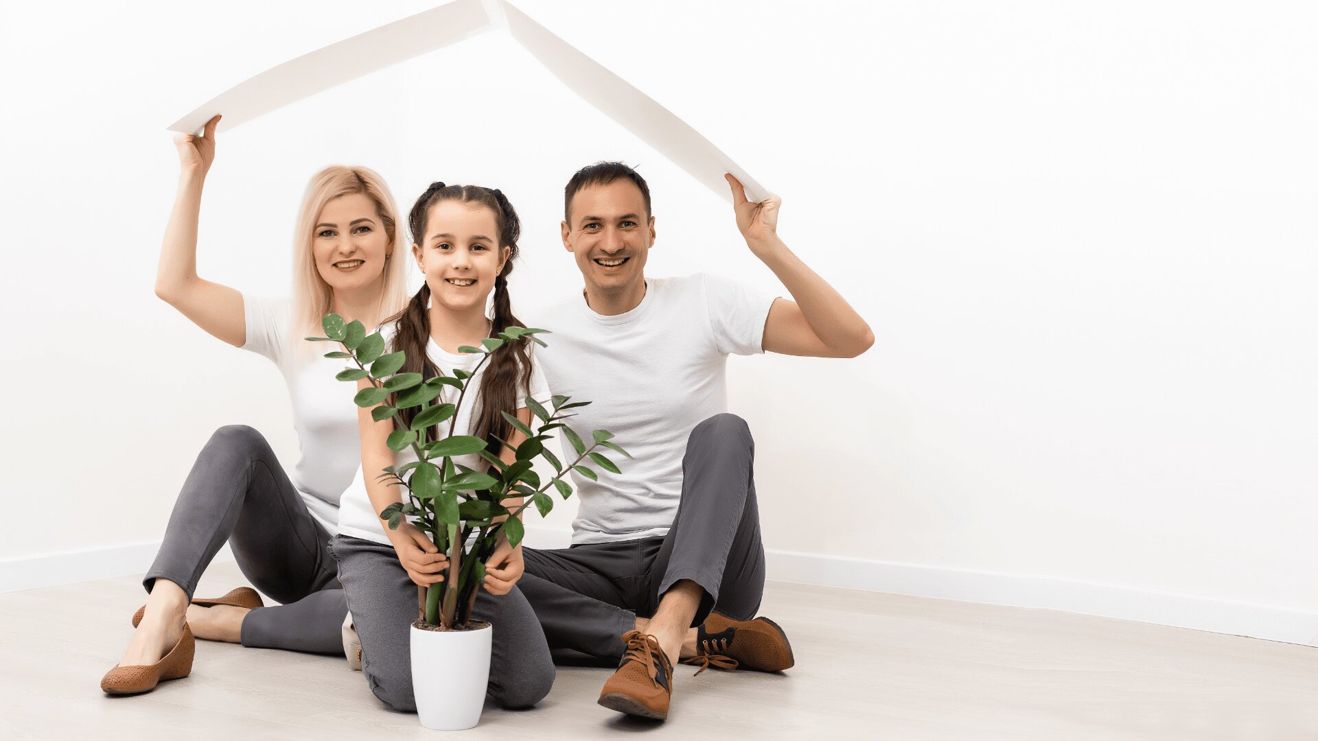 A smiling family of three sits on the floor, holding white boards above their heads like a roof. The parents are on either side of a young girl, who holds a potted plant. All are dressed in white tops and gray pants.