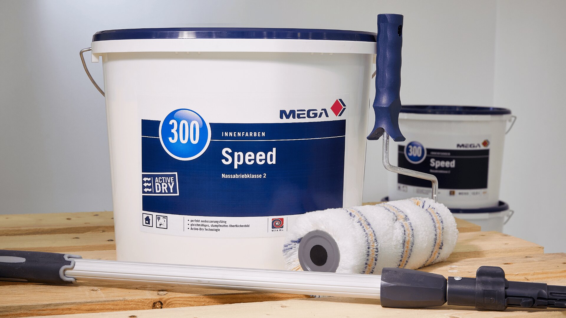 Ein großer Eimer mit MEGA 300 Speed Innenfarbe steht auf einer Holzfläche neben einer Farbrolle, einer Verlängerungsstange und einem Farbroller. Im Hintergrund ist ein weiterer ähnlicher Farbeimer zu sehen.