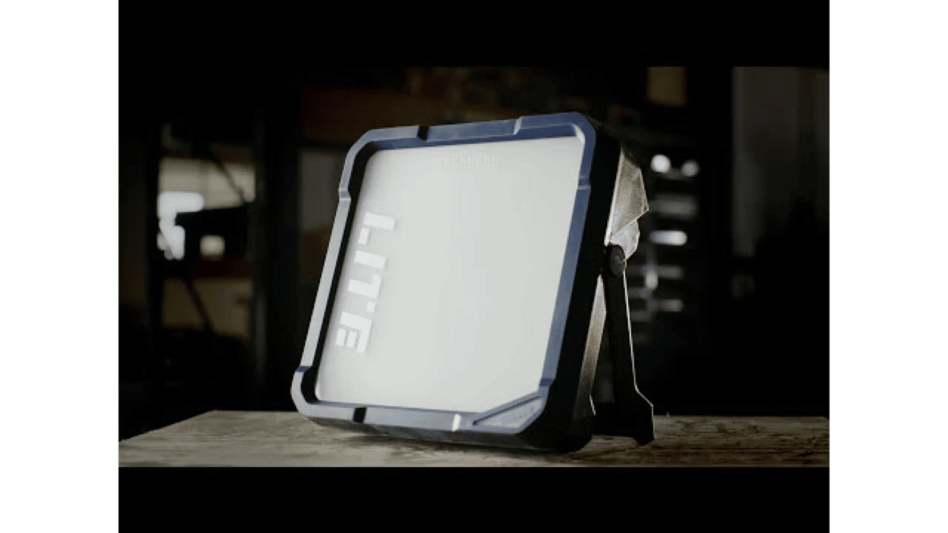 A portable, rectangular LED work light with a blue and black frame sits on a wooden surface in a dimly lit workshop. The word LITE is visible on the frosted front panel.