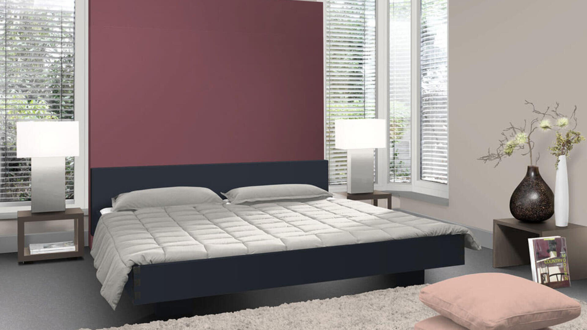 Ein modernes Schlafzimmer mit einem großen Bett in der Mitte, grauer Bettwäsche, zwei Nachttischen mit weißen Lampen, einem weichen Teppich, einem rosa Akzentkissen und einer Vase mit Zweigen auf einem niedrigen Tisch neben hohen Fenstern mit Jalousien.