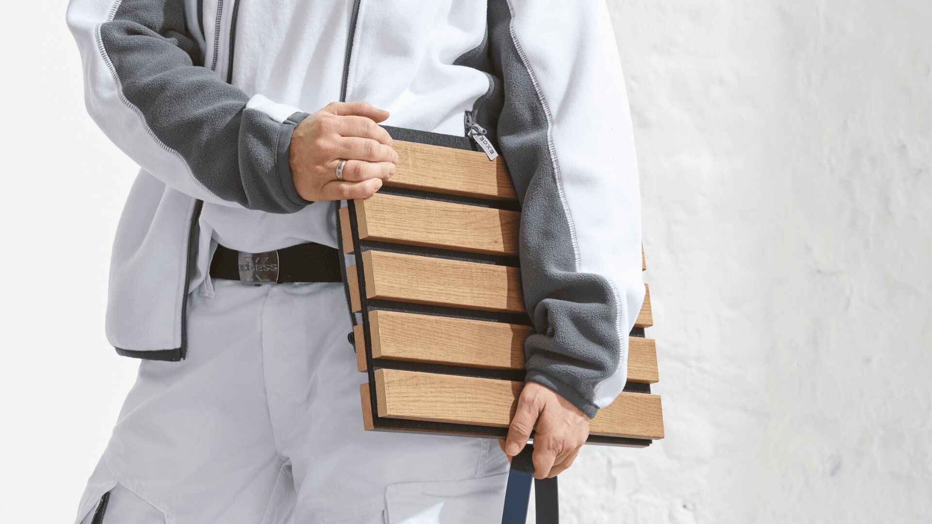 Eine Person in einer weiß-grauen Jacke hält vor einem weißen Hintergrund eine rechteckige Tasche aus Holzlatten mit horizontalen Paneelen und einem dunklen Riemen.