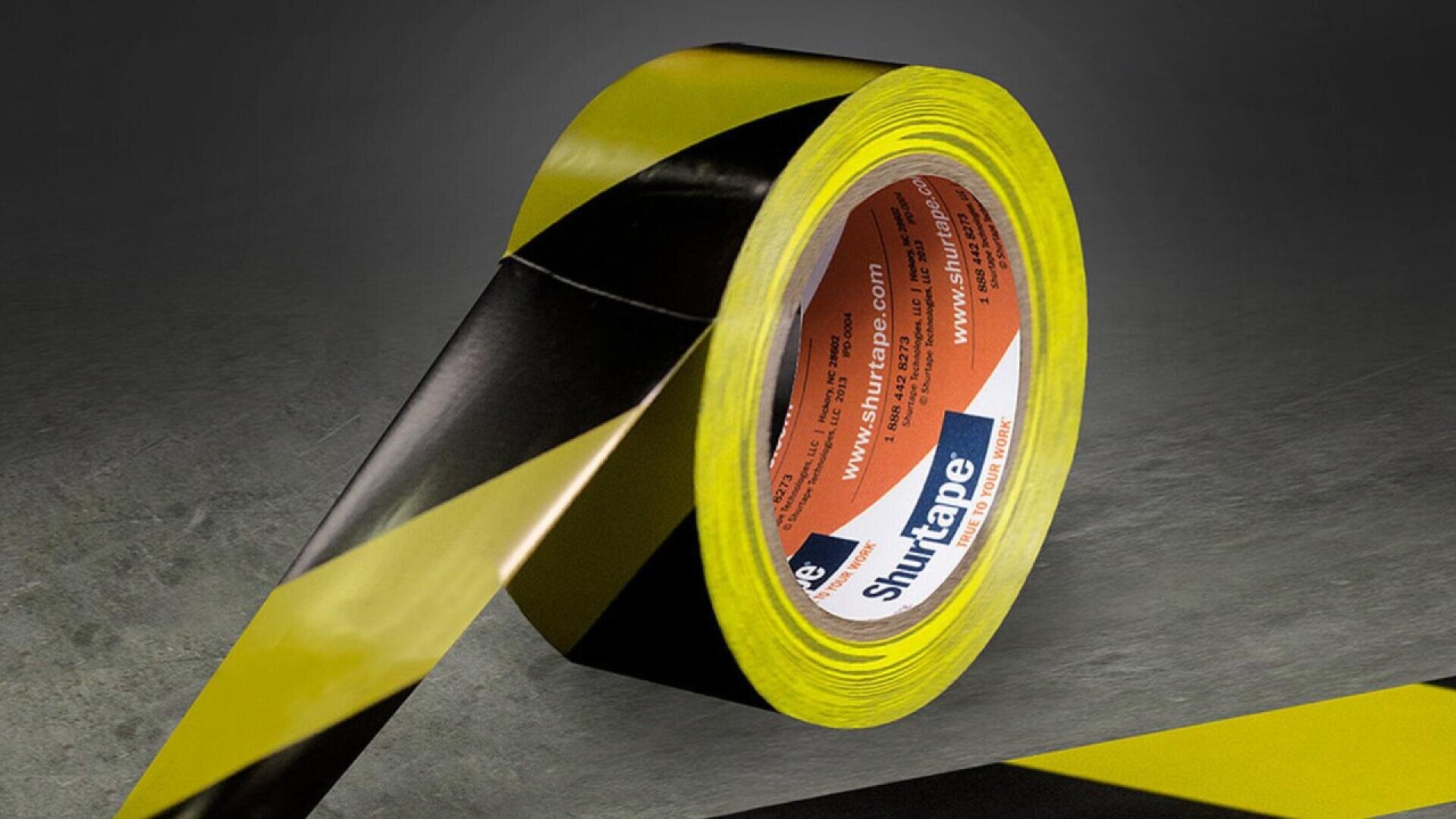 A roll of black and yellow striped adhesive tape sits on a gray surface with a strip partially unrolled. The tape is branded Shurtape and appears to be used for marking hazardous areas.