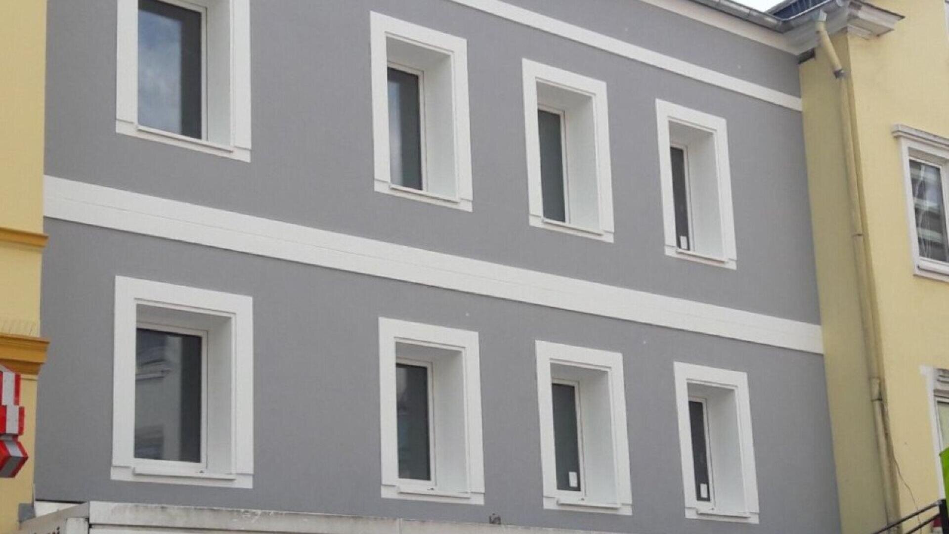 A gray and white building facade with six rectangular windows, three on the upper floor and three on the lower floor, framed in white. Yellow buildings are visible on either side.