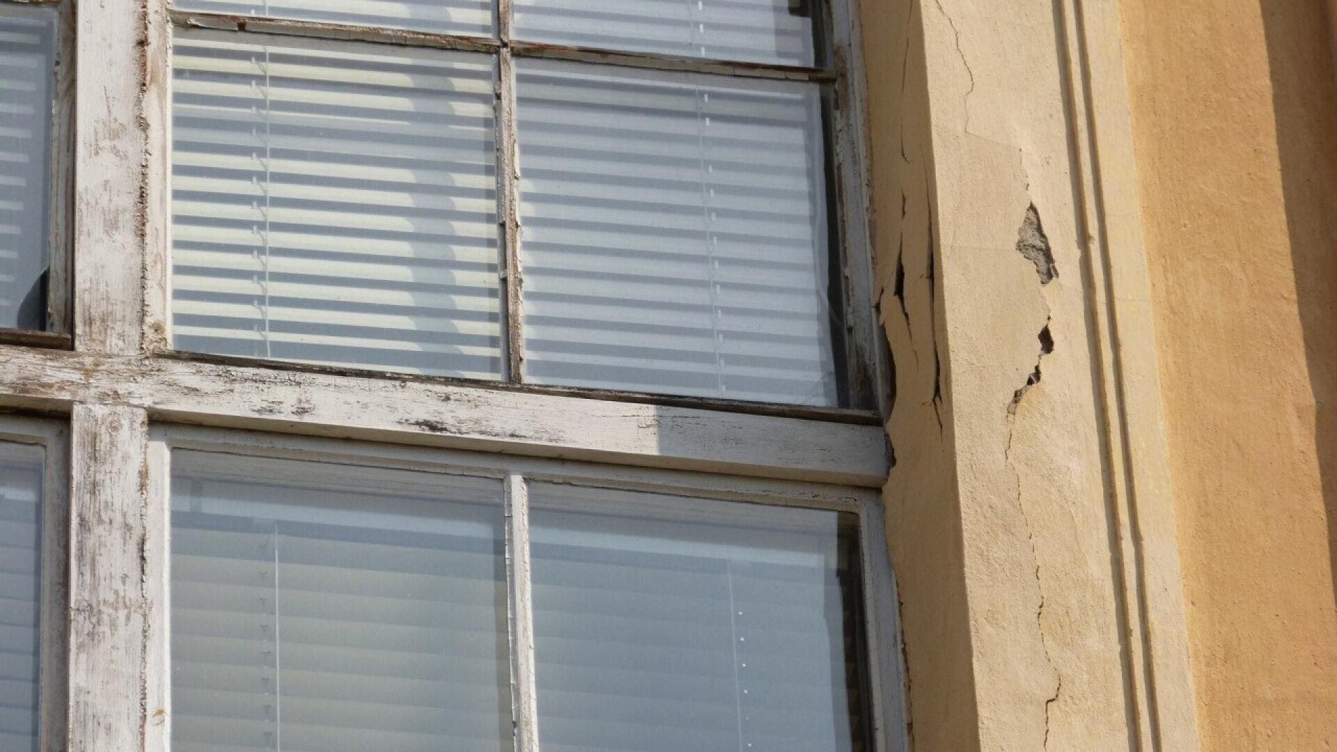 A close-up of an old window with peeling white paint on the wooden frame. The building’s yellow wall is cracked and weathered. Behind the glass, white horizontal blinds are partially closed.