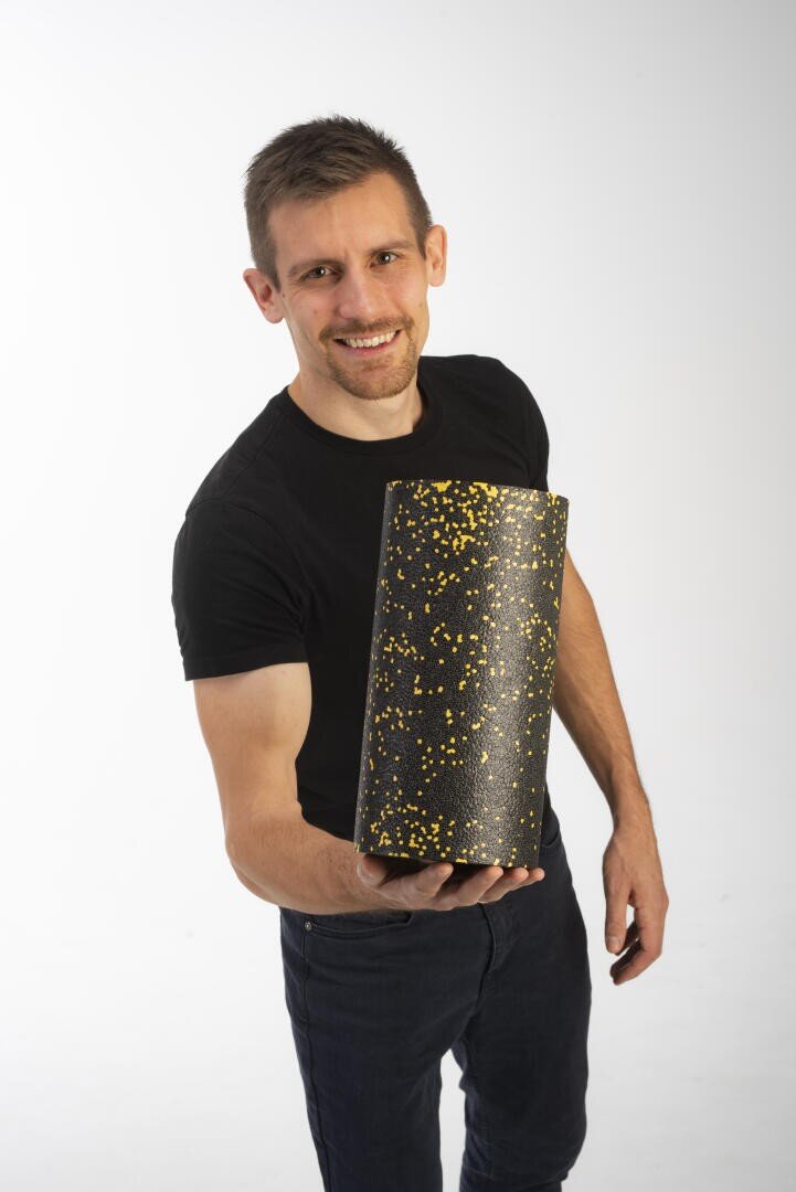 Ein lächelnder Mann in einem schwarzen T-Shirt hält vor einem schlichten weißen Hintergrund eine schwarze Schaumstoffrolle mit gelben Flecken in Richtung Kamera.
