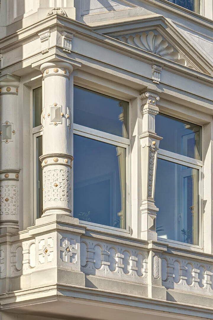 Nahaufnahme einer verzierten weißen Gebäudefassade mit großen Fenstern, dekorativen Säulen, geschnitzten Details und einem kleinen Balkon mit komplizierten Mustern und goldenen Akzenten.