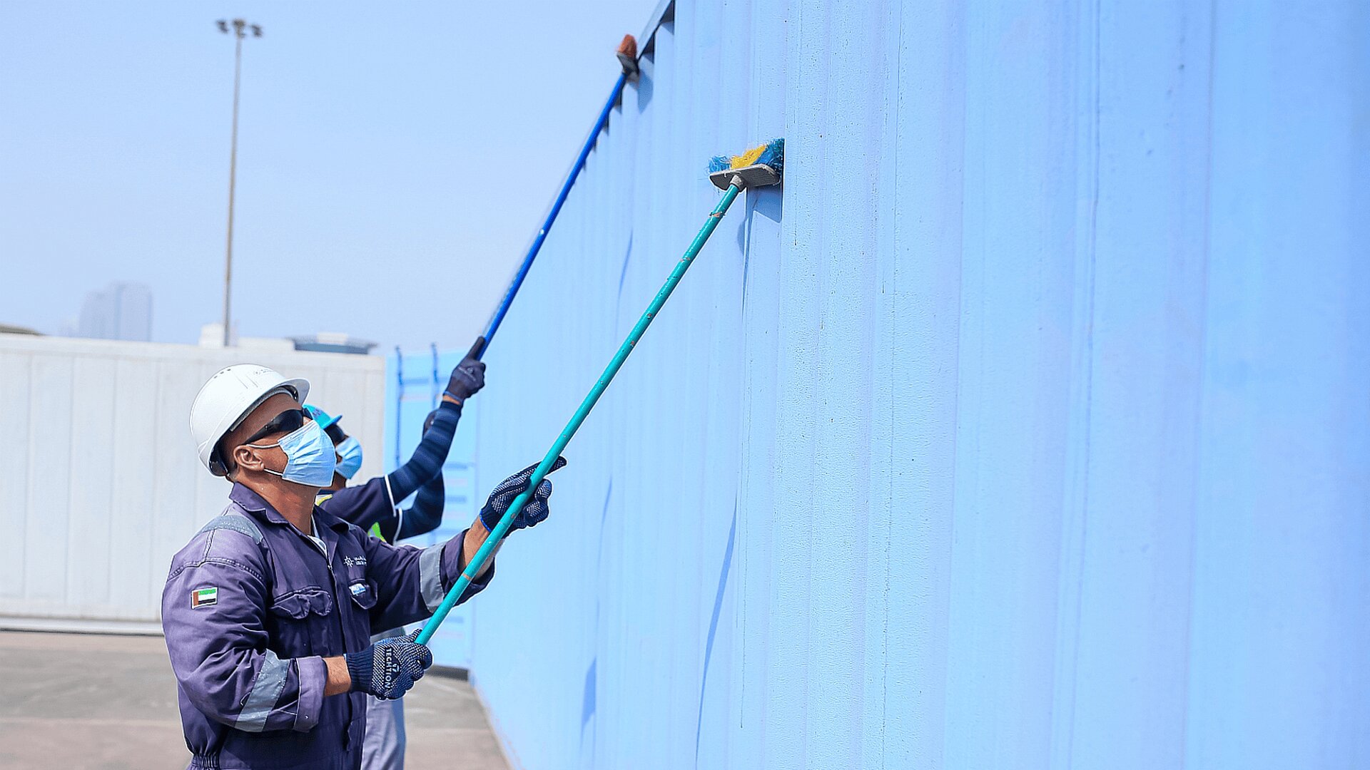 Zwei Arbeiter mit Helmen, Masken und Uniformen streichen im Freien unter klarem Himmel mit langstieligen Farbrollen eine große blaue Wand.