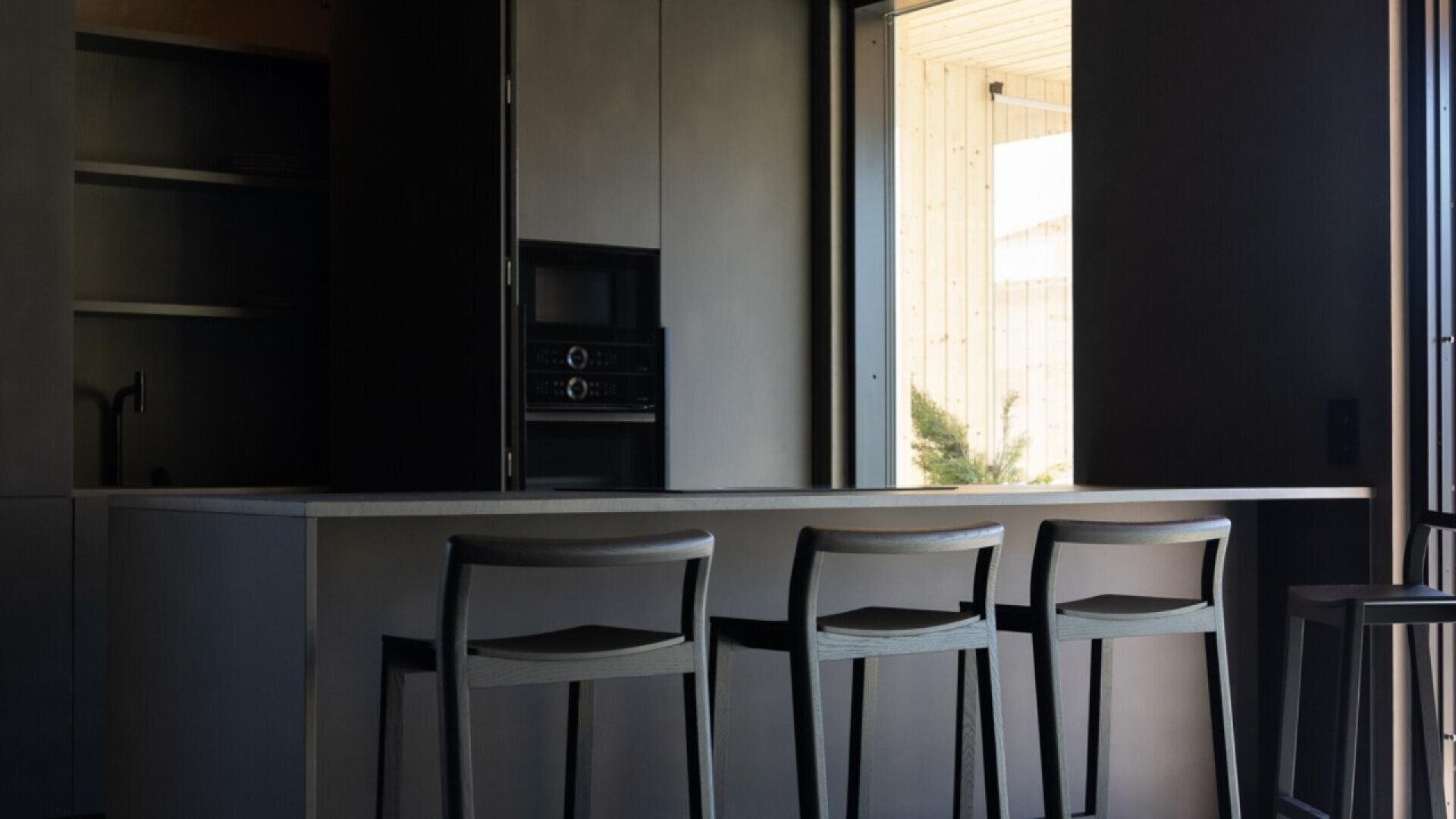 A modern kitchen with dark cabinets, an island countertop, and three high stools. Sunlight enters through a large window, partially illuminating the space.