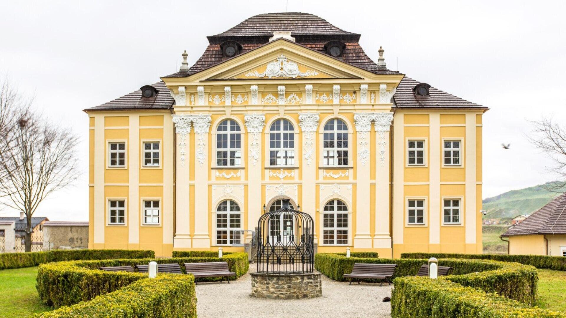 A yellow baroque-style building with white decorative details, arched windows, and a symmetrical hedge garden in front, featuring a central metal gazebo and benches along gravel paths.