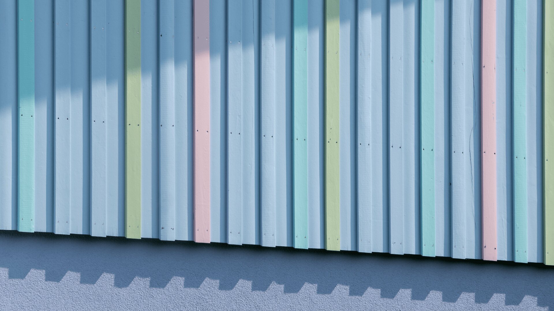 Wellblechwand mit vertikalen Pastellstreifen in Blau, Rosa, Grün und Blaugrün bemalt, wobei das Sonnenlicht eckige Schatten auf die Oberfläche im oberen und unteren Randbereich wirft.